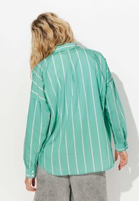 Chemise verte à rayures avec des manches longues, col, et poignets à boutons. Texture lisse avec des rayures verticales blanches. Vue arrière présentée.