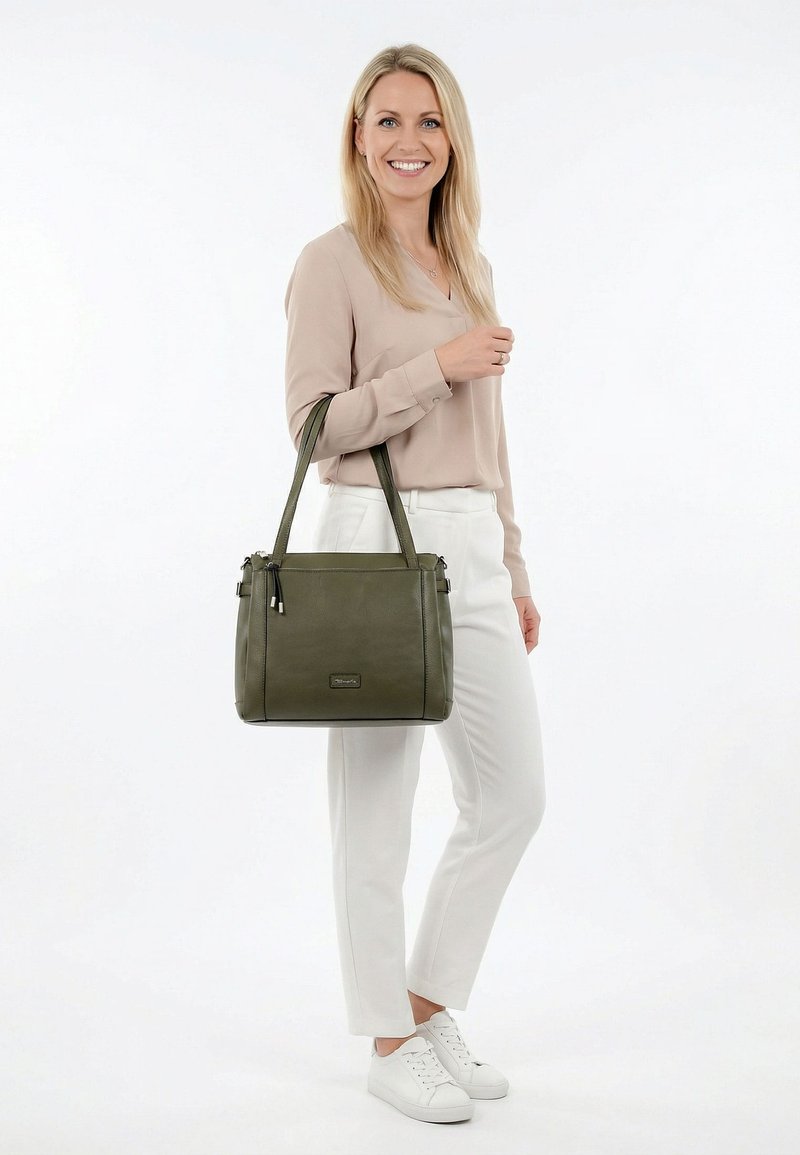 Smiling woman wearing beige blouse and white pants, holding olive green handbag over shoulder, standing on white background.
