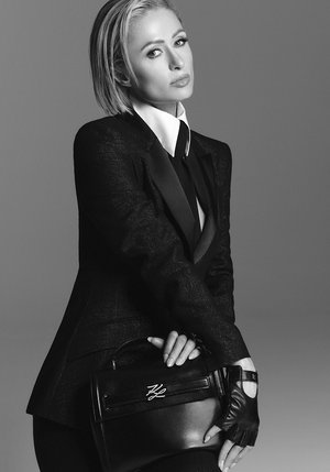 Black tailored suit with a satin lapel, white shirt with a high collar, black leather gloves, and a structured handbag with a logo.
