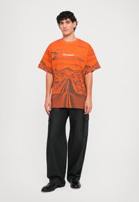 Orange grafisk t-shirt med et landskabsdesign med bjerge og en vej, parret med sorte cargo-bukser og sorte sko.