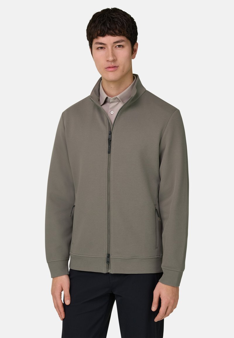 Jeune homme portant une veste grise zippée par-dessus une chemise beige et un pantalon noir, debout devant un fond uni clair.