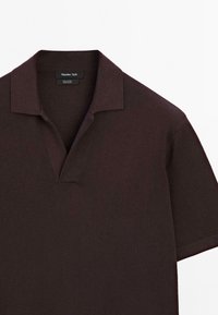 Mørkebrun kortærmet polo-shirt med et struktureret stof, der har en flad krave og en halvdækket knaplukning. Mærkatet er synligt ved halsen.