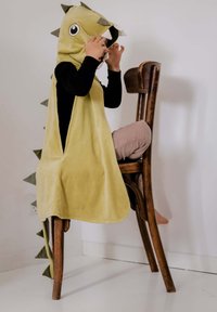 Capa temática de dinosaurio verde con capucha, tela suave y detalles en blanco. Presenta picos triangulares en la espalda. Usada por un niño sentado.