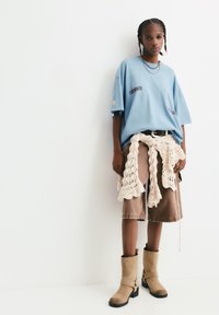 Helles blaues Oversize-T-Shirt mit dem Text "KOPENHAGEN", beigefarbene gehäkelte Accessoires an der Taille, braune Cargo-Shorts und beigefarbene High Heels.