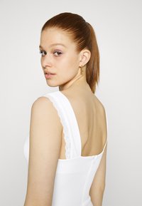 Vestido branco sem mangas com alças de ombro acentuadas por renda. Apresenta um design ajustado e um fecho de zíper nas costas discreto. Textura suave.