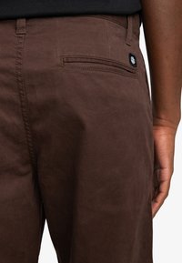 Bruine stoffen broek met een gladde textuur, achterzak met een klep en een logo-label. Het ontwerp heeft strakke lijnen en een aansluitende pasvorm.