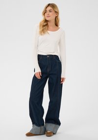 Mørkeblå jeans med vide ben og lyse denimmanchetter, med orange syninger. Kombineret med en langærmet hvid top. Blød stoftekstur.