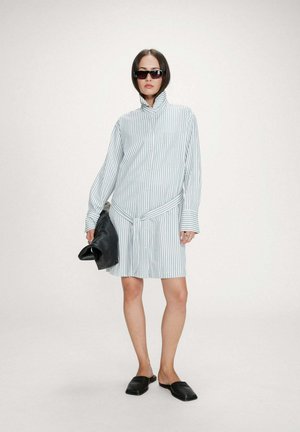 Femme portant une robe chemise à manches longues rayée bleu et blanc, des lunettes de soleil noires, tenant une pochette en cuir noire, et des chaussures noires sans lacets.