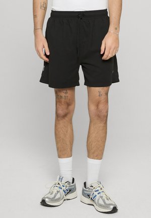 Mann iført svarte shorts med snor, hvite sokker, sølv- og blå løpesko, med synlige tatoveringer på ben og armer.