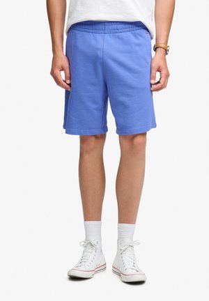 Hombre que lleva pantalones cortos deportivos azules, calcetines blancos hasta la mitad de la pantorrilla, zapatillas blancas de caña alta y una camisa blanca, de pie con las manos relajadas a los lados.