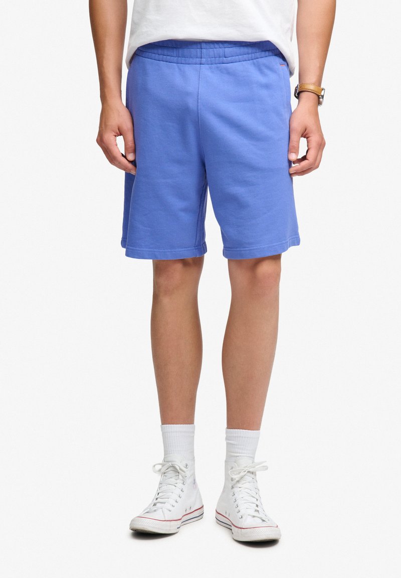Hombre que lleva pantalones cortos deportivos azules, calcetines blancos hasta la mitad de la pantorrilla, zapatillas blancas de caña alta y una camisa blanca, de pie con las manos relajadas a los lados.