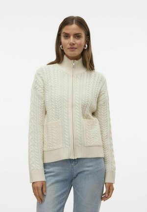 Pull zip de couleur crème avec un tricot torsadé, col haut, poignets côtelés et deux poches frontales. Tissu doux et texturé avec une finition légère.