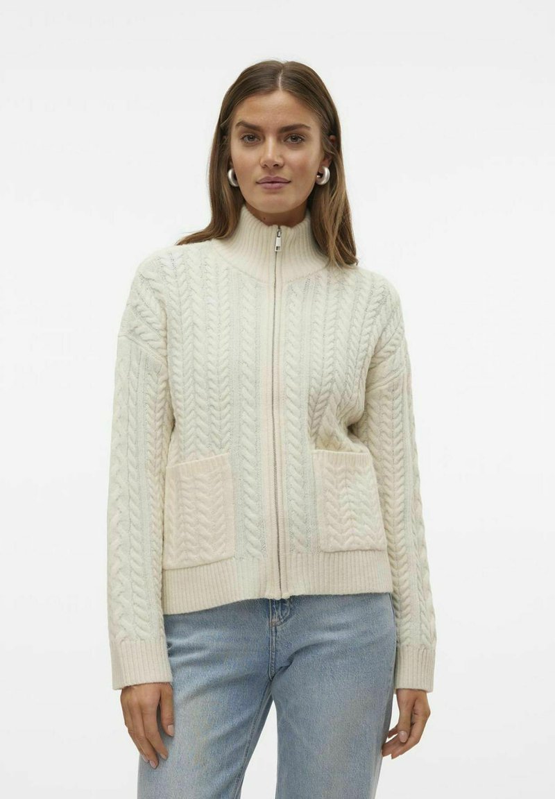 Kreminė kabelių mezgimo zip-up megztinis su aukštu apykakle, raukšlėtomis rankovėmis ir dviem priekinėmis kišenėmis. Minkšta, tekstūruota medžiaga su lengvu apdailos efektu.
