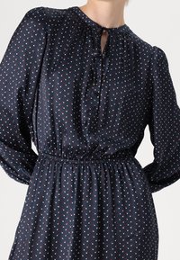 Nainen, joka käyttää tummansinistä mekkoa, jossa on pieniä punaisia ja valkoisia pilkkuja, pitkät puffihihat, napitettavassa edessä ja solmittava kaula-aukko.