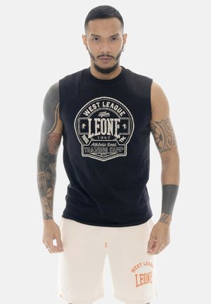 Uomo con capelli corti e tatuaggi che indossa una canotta nera e pantaloncini bianchi con scritte e loghi West League Leone.