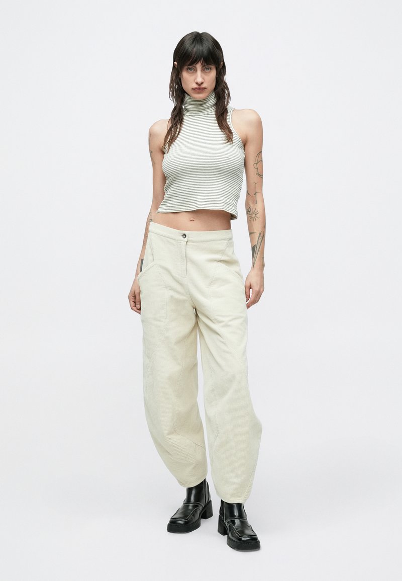 Helle beige Cordhosen mit weitem Bein und Vordertaschen, kombiniert mit einem kurz geschnittenen, grau gestreiften Rollkragenoberteil. Schwarze, hochhackige Schuhe runden den Look ab.