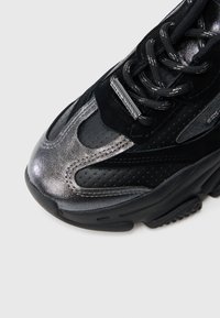 Musta urheilukenkä, jossa on rei'itetty päällinen, kiiltävät metalliset yksityiskohdat, pehmustettu kieliosa ja kuvioitu musta kumipohja.