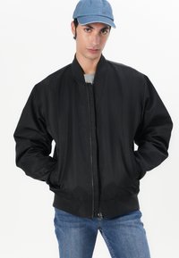 Blouson aviateur noir avec fermeture éclair à l'avant, poignets et col côtelés. Tissu lisse avec une coupe décontractée, associé à un jean bleu.