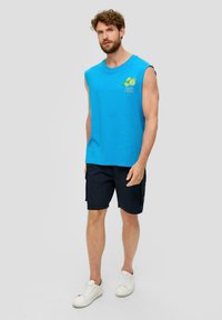 Blauwe mouwloze katoenen T-shirt met grafische print op de borst, gecombineerd met donkere shorts en witte sneakers. Eenvoudig, casual outfit ontwerp.