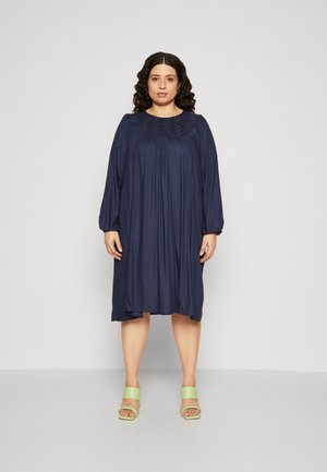 Navy blue midi dress with long puff sleeves, pleated detailing at the neckline, and small embellishments. Worn with light green block-heeled sandals.