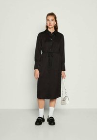 Mulher vestindo um vestido preto de manga longa com botões e cinto na cintura, meias brancas, sapatos mocassins pretos e segurando uma pequena bolsa branca.