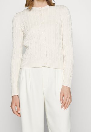 Mujer que lleva un cárdigan blanco de punto trenzado abotonado, combinado con pantalones blancos de pierna ancha y talle alto, sobre un fondo liso.