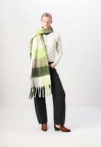 Wool scarf in green and cream stripes, fringed edges. Model wearing a fitted white top and black jeans, with brown and cream ankle boots.