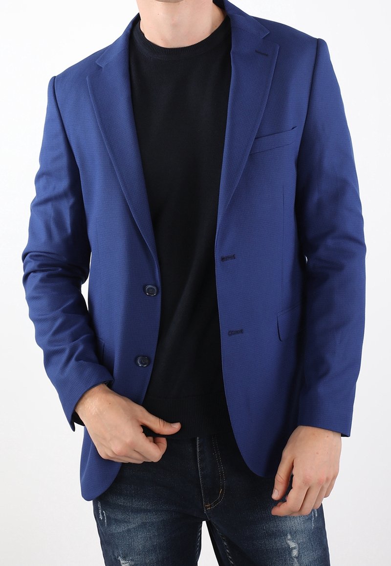 Blazer blu con texture, due bottoni, rever a pistagna e taschino, indossato sopra un maglione nero, abbinato a jeans strappati.