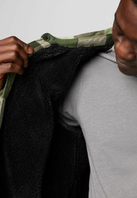 Fleece-futterte Jacke mit grün-weißem Karomuster außen, schwarzem Innenfutter und Druckknopfverschluss, die eine weiche Textur und warmes Design zeigt.