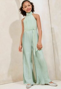 Combinaison plissée vert pâle avec un col montant et une taille à nouer. Elle présente des jambes larges fluides et des chaussures plates argentées brillantes. Texture lisse.