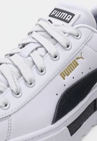 Gros plan d'une basket blanche Puma avec des lacets blancs, une bande latérale noire et le logo Puma doré sur le côté.