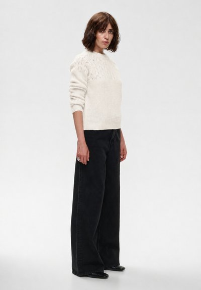 Femme aux cheveux bruns mi-longs, portant un pull en maille blanc, un pantalon large noir et des chaussures noires, debout devant un fond uni.