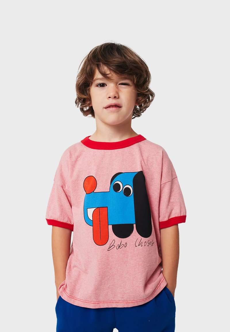 Dreng med bølget brunt hår iført en pink t-shirt med et farverigt tegneseriehundemotiv og blå bukser, stående foran en ensfarvet baggrund.