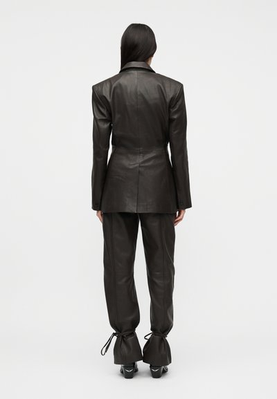 Femme aux longs cheveux foncés portant un blazer en cuir marron foncé ajusté et un pantalon en cuir assorti serré aux chevilles, debout de dos.