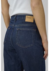 Close-up of person wearing dark blue denim jeans with a brown leather label reading "MY ESSENTIAL WARDROBE" on the waistband.