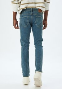 Jeans di denim blu con vestibilità dritta, design a cinque tasche e un'etichetta in pelle beige sulla vita posteriore. Sneakers bianche completano l'outfit.