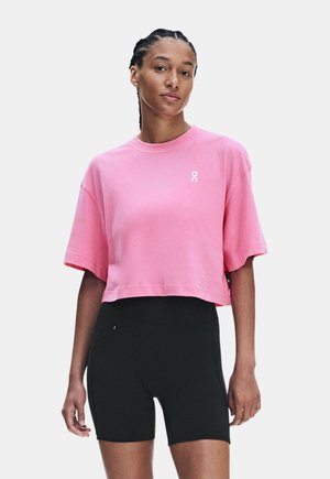 Donna con i capelli intrecciati che indossa una maglietta corta rosa e pantaloncini neri aderenti, in piedi davanti a uno sfondo chiaro e uniforme.