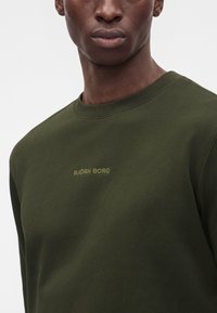 Jeune homme portant un sweat-shirt col rond vert olive avec un petit logo jaune Björn Borg sur la poitrine, sur un fond clair uni.