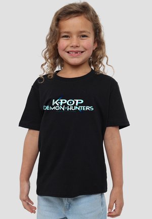 Smiling child with curly hair wearing a black "KPOP Demon Hunters" t-shirt and light blue jeans against a plain gray background.