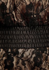 Tessuto in pizzo floreale nero con una base beige, caratterizzato da una fascia elastica nera arricciata al centro. Dettagli intricati del motivo in pizzo sono presenti.