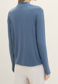 Naisten pitkähihainen paita pehmeästä sinisestä kankaasta, jossa on rento mitoitus ja minimaalinen pääntie. Sujuva pinta, jossa on hienovarainen kiilto.