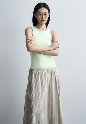 Jeune femme aux longs cheveux noirs et lunettes, debout bras croisés, portant un haut sans manches vert clair et une jupe beige rayée.