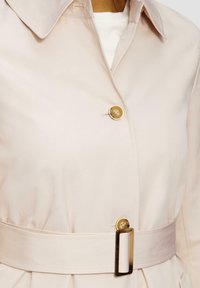 Lichtbeige trenchcoat met een knoopsluiting, grote kraag en een bijpassende riem met een rechthoekige gouden gesp en geplisseerde details.