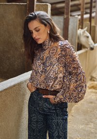 Blusa de manga larga con estampado de paisley en tonos crema y morado, con mangas abullonadas. Pantalones de cintura alta con patrón oscuro y cinturón marrón. Caballo al fondo.
