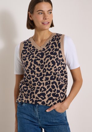 Kvinde med kort brunt hår har en leopardmønstret ærmeløs sweater over en hvid kortærmet skjorte, kombineret med blå jeans, hånd i lommen.