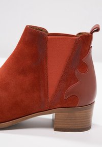 Botín de ante rojo a la altura del tobillo con paneles laterales elásticos, acentos decorativos de cuero y un tacón bajo de madera. Textura suave con elementos en contraste.