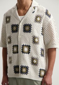 Kanten gebreid shirt in roomkleur met geometrische patronen in donkerblauw, olijf en grijs. Korte mouwen, kraag en knoopsluiting aan de voorkant.