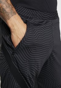 Pantalones cortos deportivos negros con un patrón texturizado de líneas onduladas. Cintura elástica y detalle de bolsillo lateral visibles. El material parece transpirable.