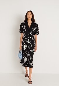 Μαύρο midi φόρεμα με φουσκωτά μανίκια, διακοσμημένο με ιριδίζουσες σπείρες, κλείσιμο με κουμπιά και γιακά με εγκοπή. Το μοντέλο κρατάει μια ανοιχτόχρωμη μπλε τσάντα.