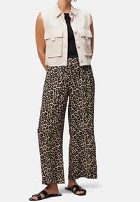 Gilet beige coupé avec poches frontales, haut noir et pantalon large à imprimé léopard. Sandales noires complètent la tenue.